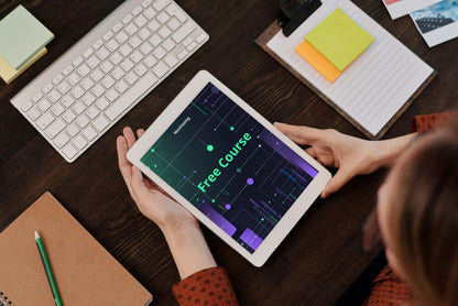 Person holding a tablet with a free course app on a desk with a keyboard and stationery.