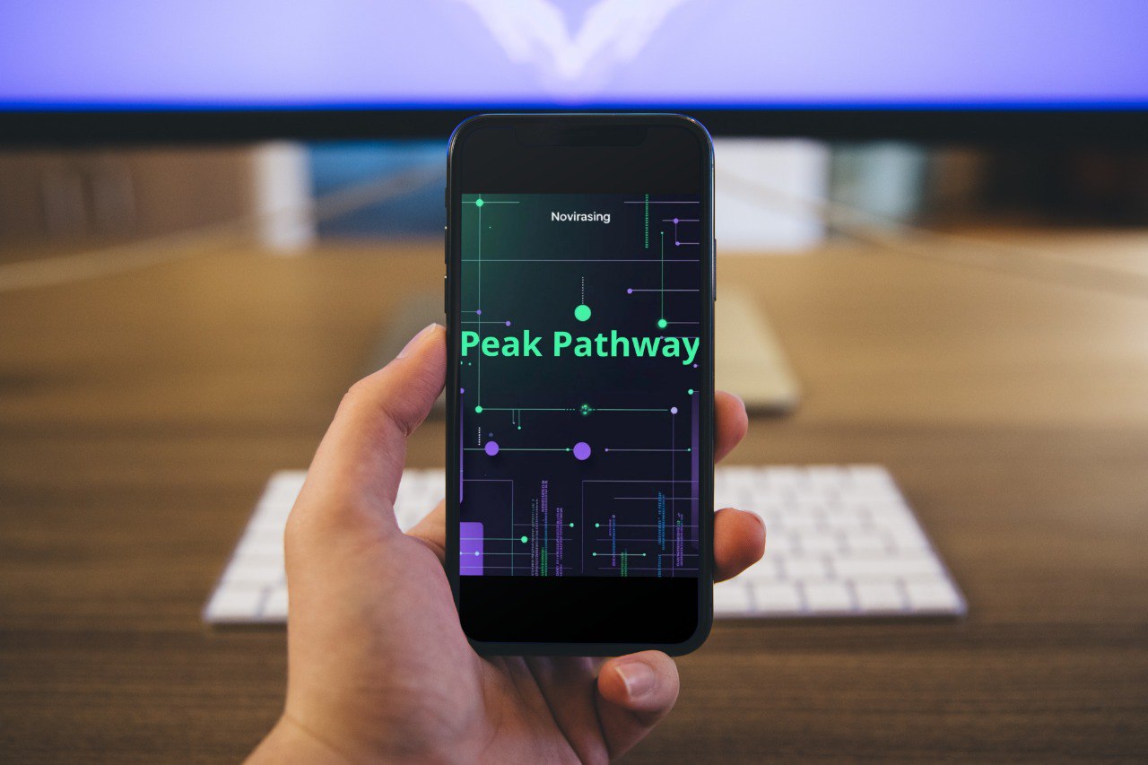 Hand holding a smartphone with 'Peak Pathway' app displayed, in front of a computer monitor and keyboard on a desk.