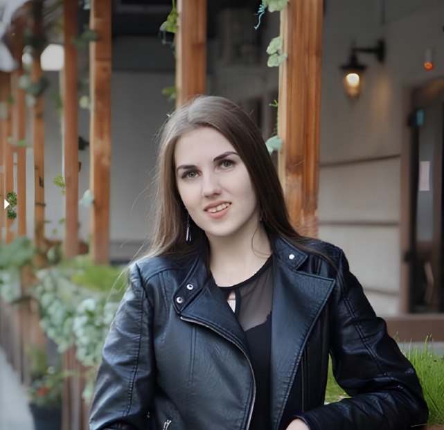 Woman wearing a black leather jacket standing outdoors with plants and wooden beams in the background